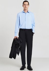 Camicia azzurra chiara button-up a maniche lunghe abbinata a pantaloni neri, tenuti insieme da una cintura nera. Giacca nera poggiata sul braccio sinistro, scarpe lucidate.