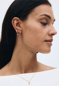 Guldörhängen med en blandning av geometriska och kurviga mönster, prydda med små ädelstenar och ett halsband med en hänge. Slät hud och axel synlig.