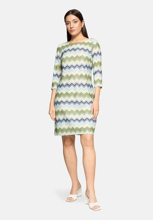 Frau steht und trägt ein knieumspielendes Kleid mit Zickzackmuster in Grün, Blau und Weiß, drei Viertel Ärmel und weiße Absatzsandalen.