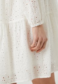 Weißes Baumwollkleid mit Spitzenstickerei, mit einem schichtweisen Design und elastischen Ärmeln. Nahaufnahme zeigt eine Hand, die auf dem Stoff ruht.