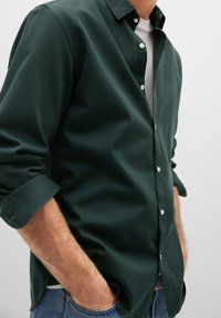 Man wearing dark green button-up shirt with sleeves rolled up and hands in the pockets of blue jeans, white undershirt visible.