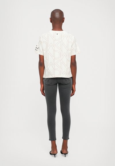 Bílý krátký rukáv tričko s béžovým textovým vzorem, spárované se šedými slim fit džíny. Modelka nosí černé vysoké muly, pohled zezadu.