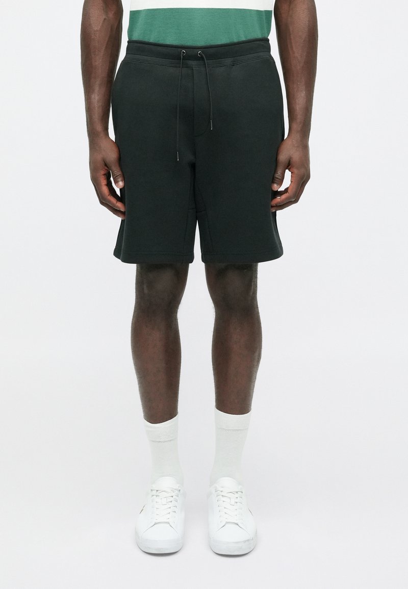 Shorts verde scuro realizzati in tessuto morbido, con vita elastica e cordoncino, tasche laterali e vestibilità rilassata. Sneakers e calzini bianchi.