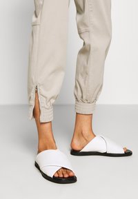 Bílé sandály na plochém černém podpatku s křížovými pásky. Design má hladký materiál a otevřenou špičku. Nosit s lehce béžovými kalhotami s ohrnutými nohavicemi.