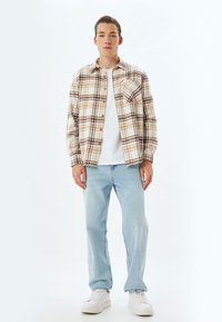 Geruite flanellen shirt in beige en bruin, gelaagd over een wit t-shirt, gecombineerd met lichtblauwe jeans en witte sneakers.