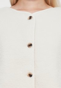 Veste en polaire blanche avec un col rond, fermeture à boutons, tissu texturé et boutons foncés contrastés. Détails de couture subtils le long des bords.
