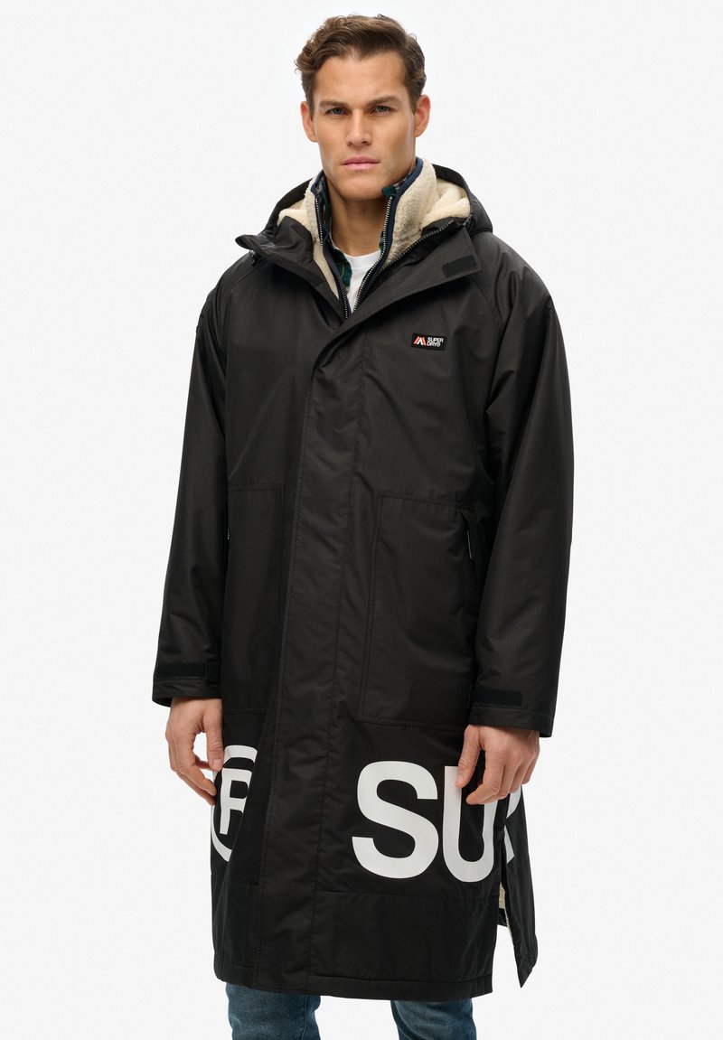 Abrigo negro impermeable con capucha, forro interior acolchado, gran logo blanco en el dobladillo y bolsillos laterales, con un diseño de corte recto.