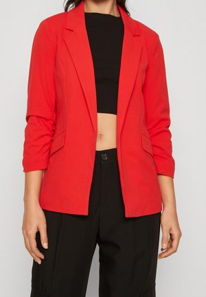 Vrouw die een felrode blazer draagt over een zwart cropped topje en zwarte broek met hoge taille, staand tegen een effen achtergrond.
