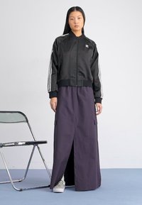Black satin bomber jacket with white stripes, paired with a long purple skirt featuring a front slit and side pockets. Checkerboard sneakers.