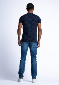 Námořnické tričko s krátkým rukávem a červenými geometrickými vzory, spolu s modrými džíny a bílými teniskami, pohled zezadu.