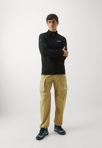 Giacca in pile nera con dettagli di cerniera gialla, pantaloni cargo beige con molte tasche e scarpe da escursionismo blu per attività all'aperto.