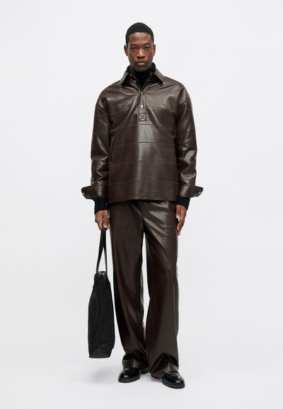 Férfi sötétbarna bőrdzseki és nadrág, fekete garbó és cipő, fekete táskát tart, fehér háttér előtt áll.