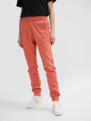 Person iført koralfarvede joggingbukser med lommer og hvide sneakers, stående mod en ensartet lys baggrund.