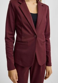 Bordeaux bleiseri yksinkertaisella nappikiinnityksellä, hapsureunuksilla ja etutaskuilla. Valmistettu sileästä kankaasta, istuva malli, yhdistettynä samasta kankaasta valmistettuihin housuihin.