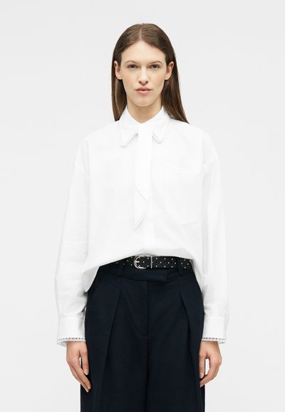 Femme aux cheveux bruns raides portant une chemise blanche à manches longues avec des bordures en dentelle et un pantalon noir taille haute avec une ceinture cloutée.