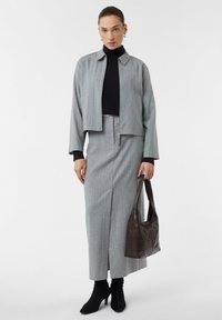 Frau in einem grauen Nadelstreifenblazer und passendem Rock, schwarzem Rollkragenpullover, schwarzen Ankle Boots und hält eine dunkelbraune Schultertasche.