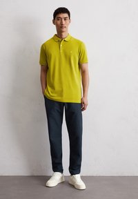 Limegrünes Poloshirt mit Kragen, kurzen Ärmeln und dezentem Logo; kombiniert mit dunkelblauen Hosen und weißen Sneakern. Einfaches, tailliertes Design.