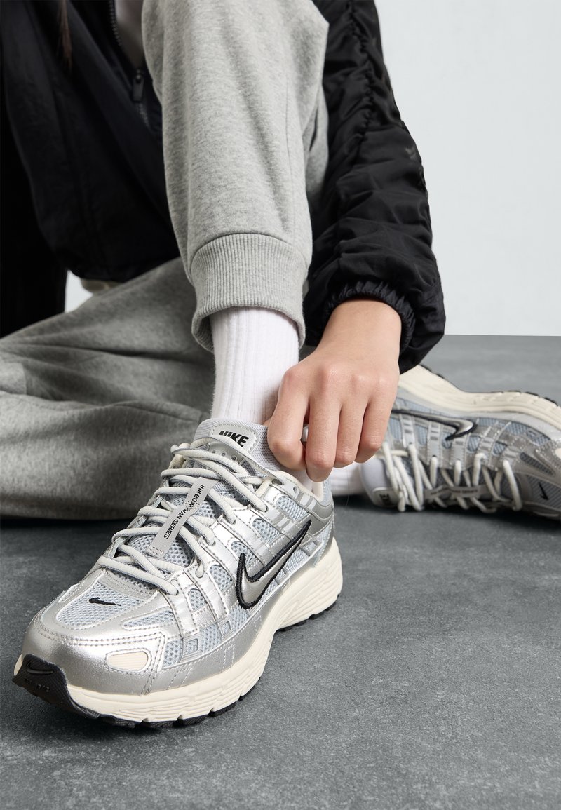 Τα αθλητικά παπούτσια Nike διαθέτουν ασημί συνθετικό πάνω μέρος με λεπτομέρειες από διχτυωτό υλικό, μαύρες λεπτομέρειες και χοντρή λευκή σόλα. Το άτομο ρυθμίζει τα κορδόνια.