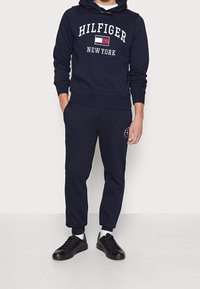 Sudadera azul marino con texto y logo en blanco "HILFIGER NEW YORK", combinada con joggers ajustados a juego y zapatillas negras.