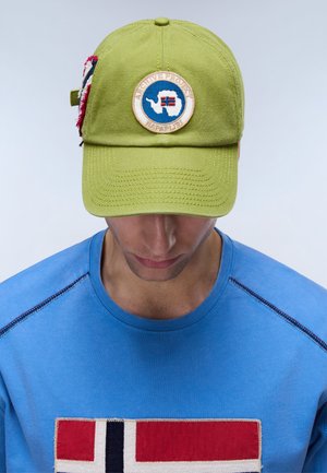 Man draagt een lichtgroene pet met een ronde patch waarop Antarctica en een vlag te zien zijn, en een blauw shirt met een groot vlagontwerp op de borst.