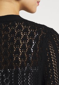 Cardigan nero lavorato a maglia con motivo a tessitura aperta, caratterizzato da un design testurizzato e un collo rotondo, che mette in risalto i dettagli dei punti intricati.
