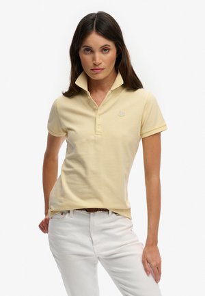 Mujer con una camisa polo de color amarillo pálido con el cuello levantado y pantalones blancos, de pie con una mano en el bolsillo contra un fondo liso.