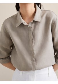 Blouse en lin gris clair avec col à revers, fermeture boutonnée et manches retroussées, présentant une texture douce et une coupe décontractée.