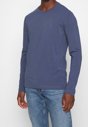 Mężczyzna ubrany w prostą, ciemnoniebieską koszulę z długim rękawem i małym logo Tommy Hilfiger na piersi oraz jasnoniebieskie jeansy.