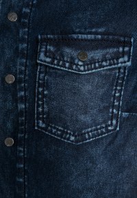 Close-up of dark blue denim jacket pocket with flap and metal snap button, showing detailed stitching and fabric texture.