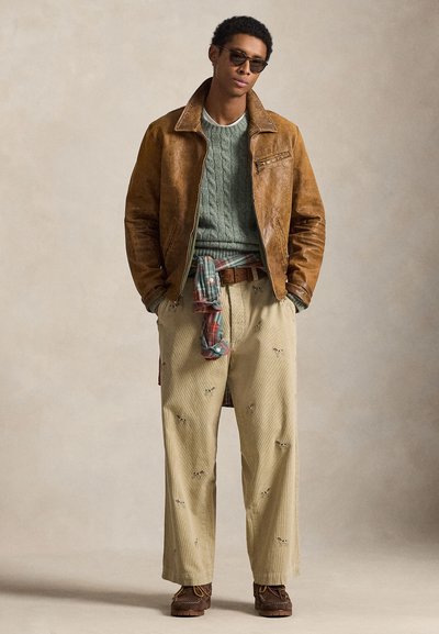 Καφέ δερμάτινο μπουφάν πάνω από πράσινο πουλόβερ με πλεκτό σχέδιο, μπεζ κεντημένο παντελόνι και καφέ παπούτσια. Αξεσουάρ περιλαμβάνουν γυαλιά ηλίου και καρό πουκάμισο δεμένο στη μέση.