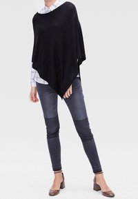 Poncho noir avec une silhouette ample et drapée, associé à un jean en denim foncé avec un design en blocs de couleurs. Une paire de chaussures à talons marron complète la tenue.