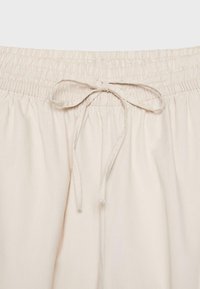 Taille élastique avec un nœud noué sur un tissu beige clair, montrant des coutures plissées et un matériau lisse sous la taille.
