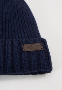 Navy blue ribbed knit hat with a brown leather tag embossed with "Barbour ENGLAND." Features a thick, textured fabric for warmth.