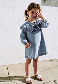 Denimmekko, jossa on vaaleansininen sävy, varustettuna laskostetulla brodeeratulla kauluksella ja pitkillä hihoilla. Yhdistettynä beigeihin sandaaleihin, seisoo kivipinnalla.