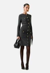 Vestito grigio in maglia con maniche lunghe, bottoni dorati, due tasche frontali e cintura abbinata. Indossato con collant neri e tacchi a punta.