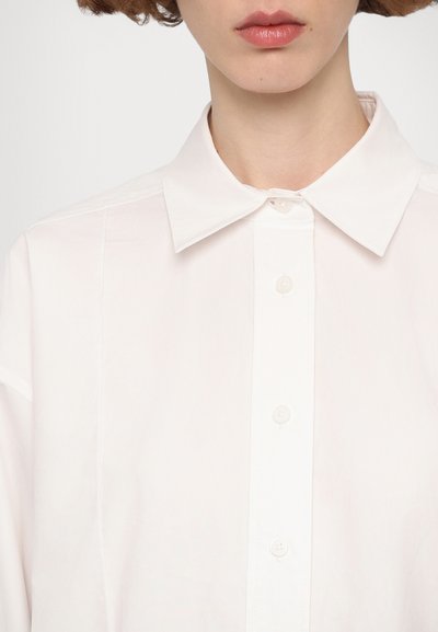 Camicia di cotone bianca con colletto classico, chiusura frontale a bottoni e vestibilità rilassata. Tessuto liscio con maniche corte.