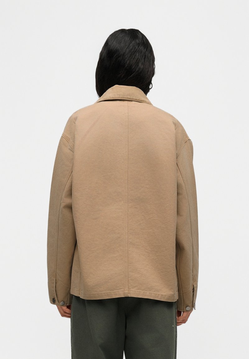 Persoon met lang zwart haar draagt een losse beige jas met een kraag, staand met de rug naar de camera tegen een effen witte achtergrond.