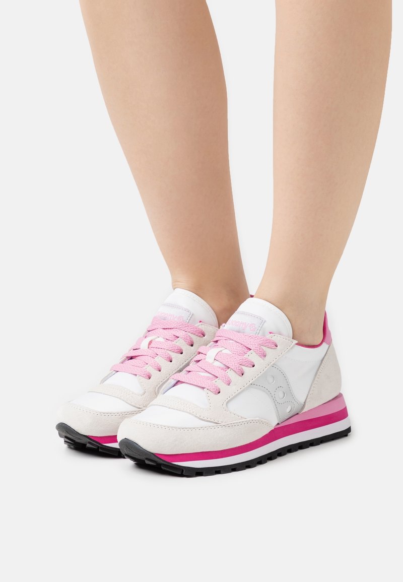 Pieds portant des baskets blanches et beige clair avec des lacets roses et des semelles rayées roses sur un fond clair.