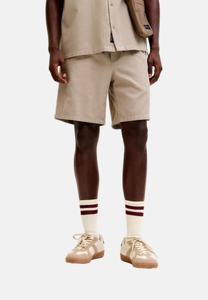 Hombre vistiendo pantalones cortos beige y zapatillas a juego, calcetines blancos con dos rayas granate, y una camisa beige, de pie con las manos relajadas.