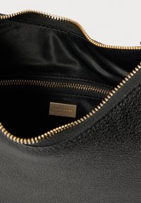 Εσωτερικό μαύρης δερμάτινης τσάντας με λεία υφή, που διαθέτει χρυσό φερμουάρ και μικρή μεταλλική ετικέτα της μάρκας.