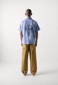 Camicia azzurro chiaro a maniche corte con logo "Detroit" sul retro, abbinata a pantaloni cargo beige a vestibilità ampia con tasche laterali e ricami.