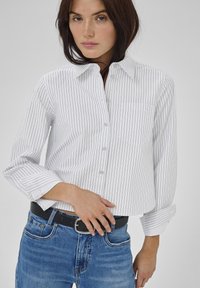 Camicia bianca con bottoni, a righe verticali nere sottili, colletto classico, taschino sul petto e maniche arrotolate, abbinata a jeans blu.