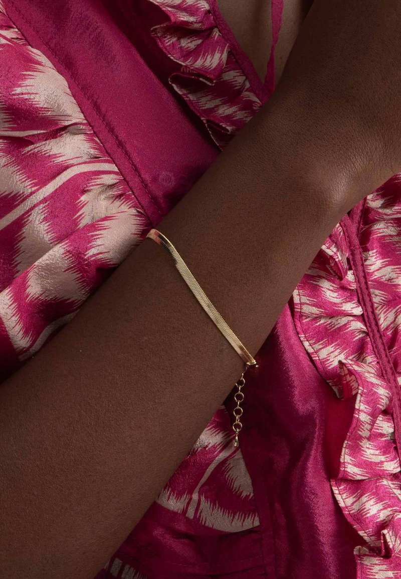 Guldarmband med en texturerad, avlång rektangulär design och kedjestängning, bärs på en mörk hudton, mot en rosa mönstrad tyg.