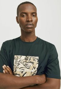 Camiseta de color verde oscuro con mangas cortas, que presenta un estampado gráfico en crema y el texto "Originals Studio" en una fuente en negrita en la parte frontal.