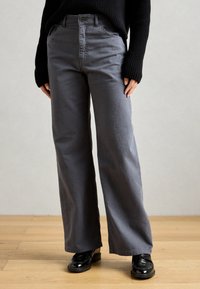 Személy széles szárú sötétszürke nadrágban, fekete, hosszú ujjú kötött pulóverben és fényes fekete loaferekben áll egy fa padlón.