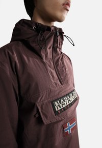 Giacca a pullover bordeaux con colletto alto, dotata di una tasca frontale, patch con logo e un accento della bandiera norvegese. Tessuto leggero e testurizzato.