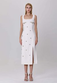 Femme blonde se tient debout, portant une robe midi blanche sans manches ornée de fleurs dorées et avec une fente à l'avant, associée à des sandales à talons blancs avec une bride à la cheville.