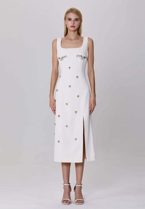 Femme blonde se tient debout, portant une robe midi blanche sans manches ornée de fleurs dorées et avec une fente à l'avant, associée à des sandales à talons blancs avec une bride à la cheville.