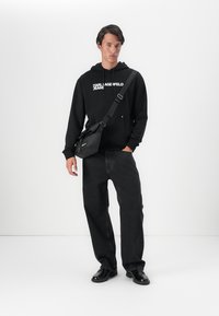 Čierna mikina s kapucňou a logom "Karl Lagerfeld Jeans", s tmavými džínsami a čiernymi topánkami, a malou čiernou kabelkou cez rameno. Jednoduchý dizajn.