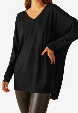 Femme portant un haut noir ample à manches longues avec col en V, épaules tombantes et leggings sombres brillants et ajustés sur fond blanc.